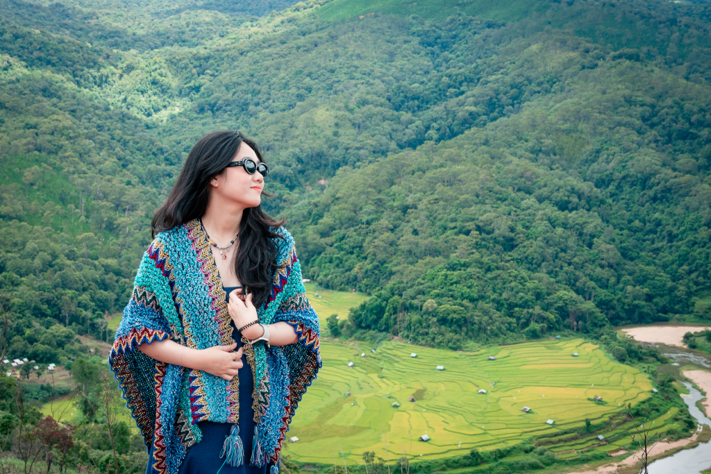 cô gái chụp ảnh tại Kon Tu Rằng với view trên cao ngắm núi rừng xung quanh, dưới thung lũng là ruộng bậc thang được bao bọc bởi con suối, xa xa bên trái là một con thác, du khách có thể tham gia tour photo tour măng đen để trải nghiệm